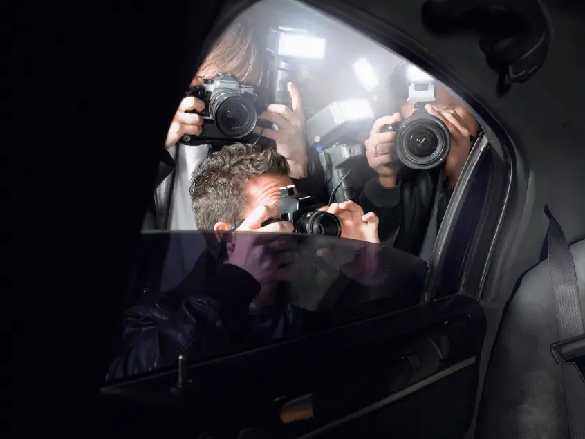 Three photographers holding cameras peering into a celebrity's car window, aiming their lenses at someone inside under dim lighting.