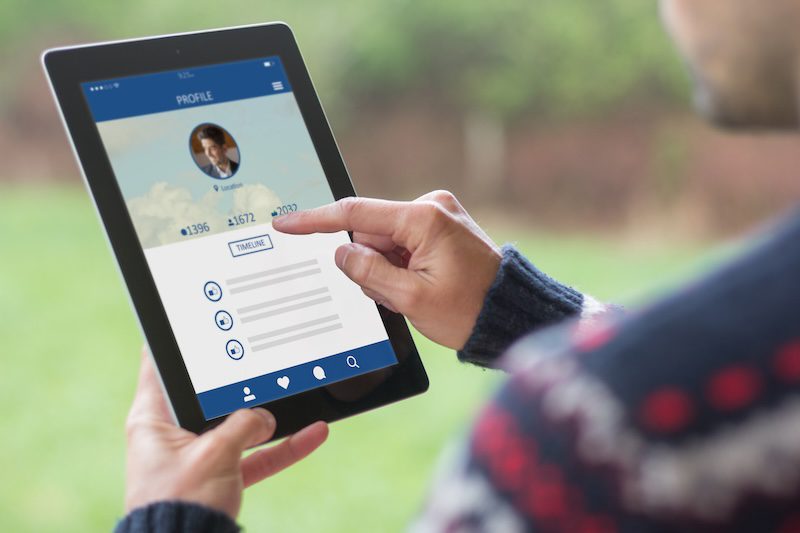 A person is holding a black tablet displaying a social media profile page. The screen shows a profile picture, follower statistics, and icons for various actions. The person, clad in a dark sweater with a patterned design, uses their index finger to navigate the screen while pondering how to remove pictures from Google Images.