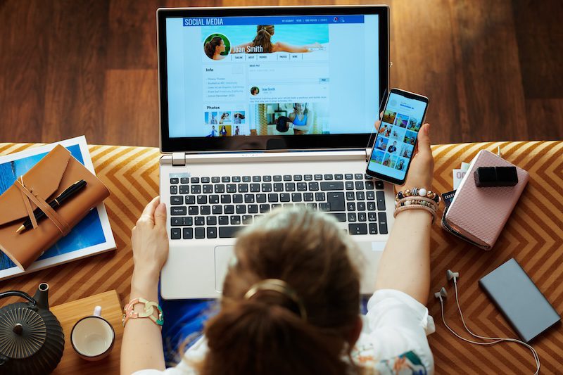 A person sits at a desk with a laptop and smartphone, browsing a social media profile. The desk holds various items, including a notebook, pen, potted plant, and earbuds. As they navigate the profile page with the header "SOCIAL MEDIA" and a name displayed, they're contemplating removing pictures from Google Images.