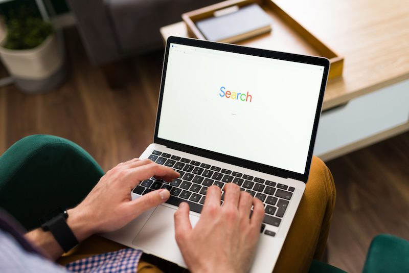 Person seated using a laptop with a search engine displayed on the screen, with the word "Search" shown in colorful letters. 