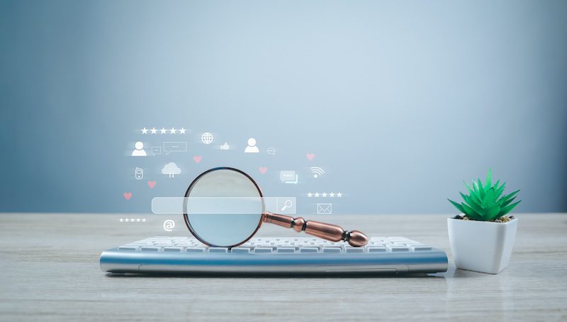 A silver keyboard sits on a light wood surface with a small potted succulent plant to the right. A magnifying glass is placed on the keyboard, with digital icons of search bars, emails, and social media symbols floating above it, symbolizing online search and removing pictures from Google Images.