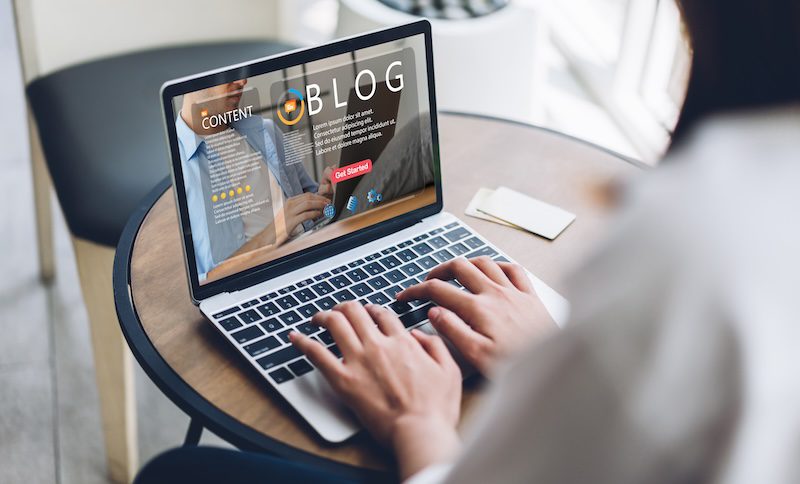 A person is typing on a laptop at a round table, creating or editing a blog. The laptop screen displays the words "CONTENT BLOG" with a "Get Started" button. Nearby, a cup and a business card rest on the table, while they mull over removing pictures from Google Images to enhance their blog's originality.