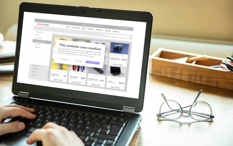 A person using a laptop displaying an online shopping site with a cookie consent popup pauses to adjust glasses resting on the desk next to a pencil holder. It's as if time were in a LexisNexis freeze, capturing this moment of digital browsing and quiet reflection.