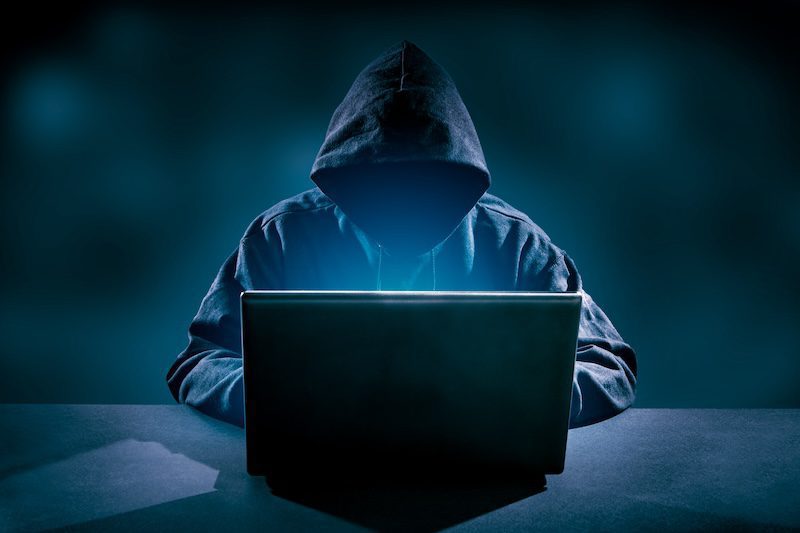 A person wearing a dark hoodie is sitting at a table, using a laptop with LexisNexis open. The room is dimly lit, casting a mysterious glow on the screen and the hooded figure, frozen in concentration.