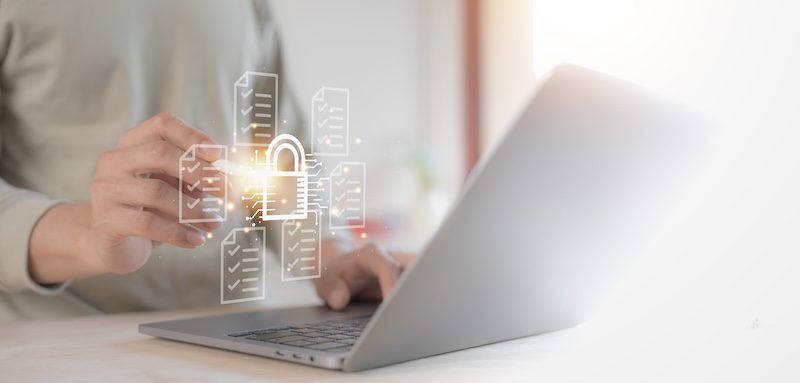 A person is using a laptop with their left hand on the keyboard. Their right hand interacts with a holographic display featuring a glowing padlock symbol and surrounding file icons, suggesting executive privacy and data protection.