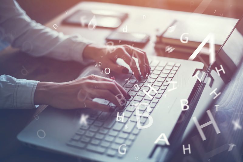 A person typing on a laptop keyboard with floating alphabetical letters overlaid in the foreground. The scene suggests concepts of digital communication, data entry, or writing. The person's hands are visible, with a stack of books and a notepad in the background, perhaps researching how to push negative search results down.