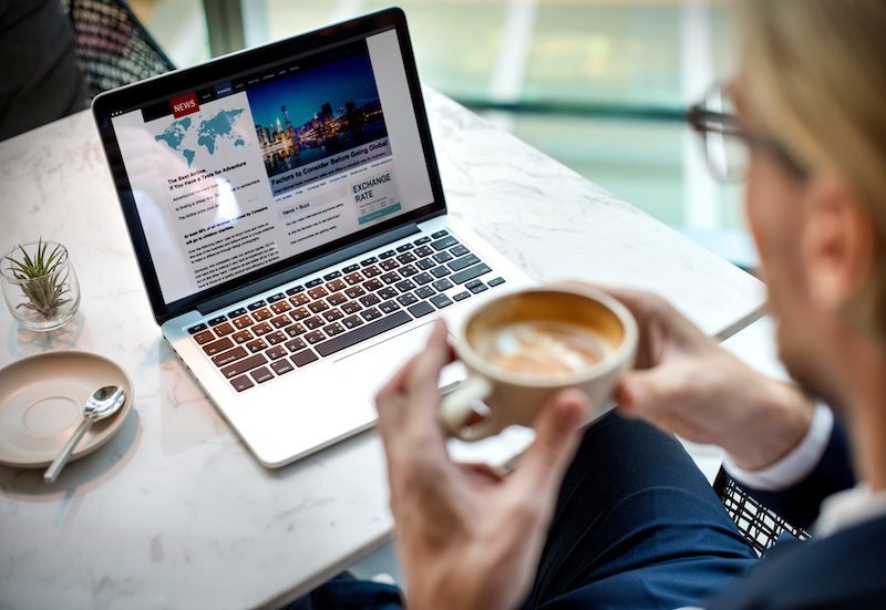 A person holding a coffee cup sits at a table with a laptop displaying a news website. The background of the laptop screen shows an article titled "How to Push Negative Search Results Down," featuring a world map and a cityscape image. A small plant and an empty saucer are also on the table.