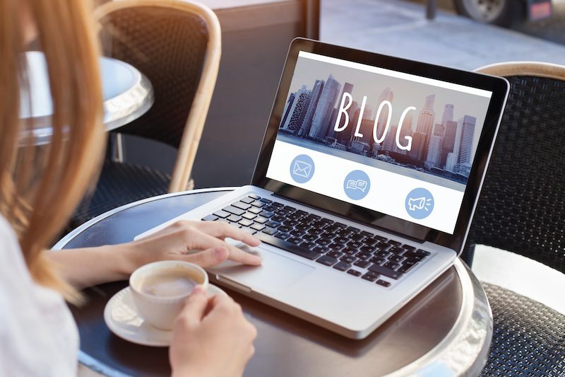 A person is sitting at a round table with a cup of coffee in hand, using a laptop. The laptop screen displays the word "BLOG" with a city skyline in the background and three icons below the text. The setting appears to be a casual, outdoor café where they're researching how to push negative search results down.