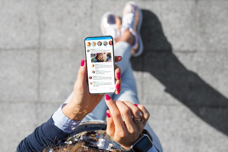 A person with pink nail polish holds a smartphone while browsing a social media app, possibly searching for how to push negative search results down. The individual is wearing a blue long-sleeve shirt and light jeans, standing on a paved surface. Only their hands and legs are visible in the frame.