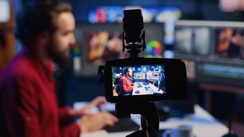 A man with a beard is sitting at a desk with multiple monitors, typing on a keyboard. The scene is captured in the viewfinder of a camera, emphasizing his focus on learning how to push negative search results down. The background reveals a blurry, colorful workspace.