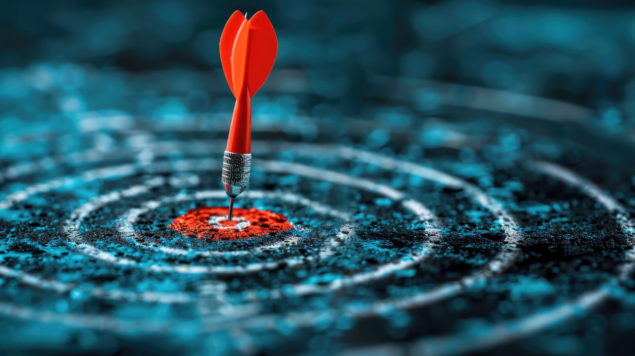 A red dart is perfectly centered on a blue and black dartboard, hitting the bullseye. The background is blurred, emphasizing the dart and the precise hit on the target.