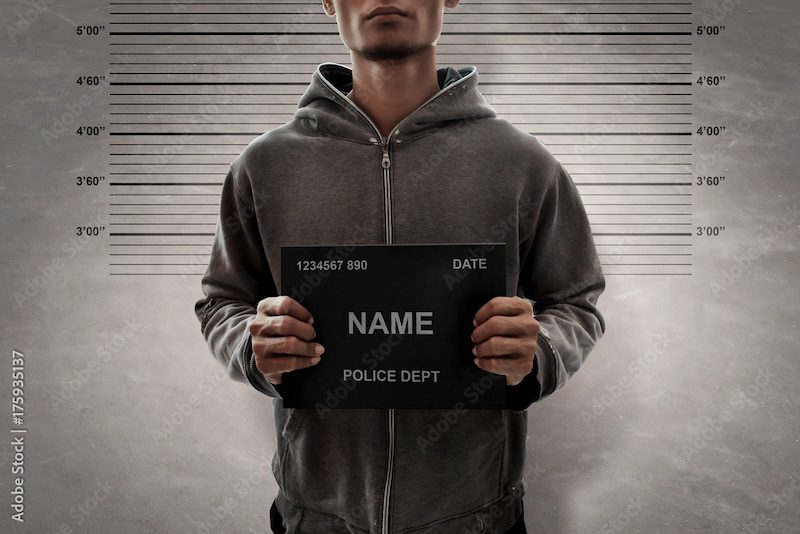 A man in a hoodie stands against a height chart, holding a police placard that reads "NAME" and "POLICE DEPT." His face is partially visible. For those wondering how to get someone's mugshot, this scene captures the essence against a backdrop of gray, textured walls with height markers.