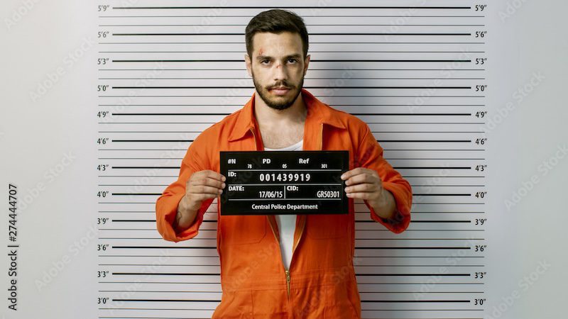 A man in an orange prison jumpsuit stands against a height chart, holding a black and white identification board. The board displays details like a code, date, and "Central Police Department," reminiscent of how to get someone's mugshot for official records.