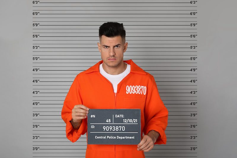 A man in an orange prison jumpsuit stands solemnly before a height chart, clutching a sign with his ID number and arrest date. With short dark hair and a serious expression, he looks like someone whose mugshot you might find when searching how to get someone's mugshot online.