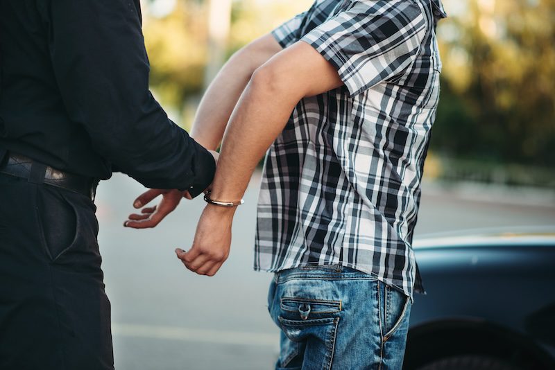 A person in a black shirt places handcuffs on another wearing a plaid shirt and jeans, standing beside a car on the street. With blurred greenery in the background, it's a scene that might prompt one to wonder how to get someone's mugshot from such an arrest scenario.