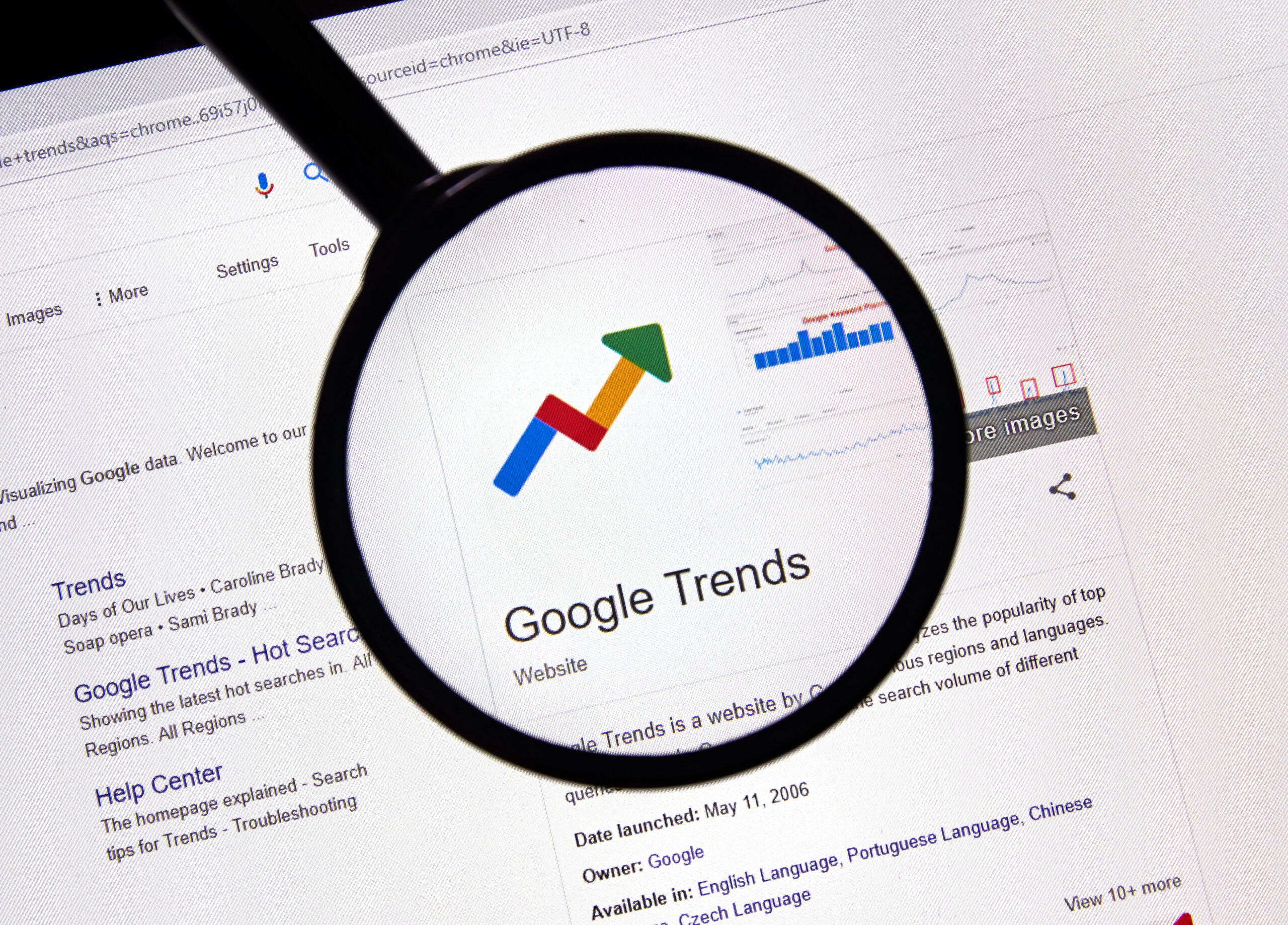 A magnifying glass focuses on the Google Trends website, with its logo featuring a colorful upward graph. The background shows a web page displaying search results related to Google Trends.