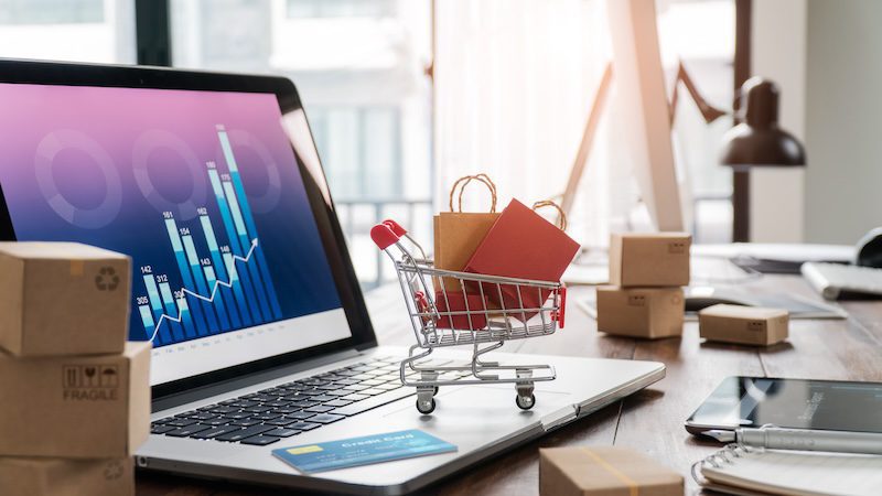 A small shopping cart with bags sits on an open laptop displaying a bar chart, surrounded by cardboard boxes and a smartphone on a wooden desk. Sunlight filters through a window in the background, suggesting an e-commerce or online business theme.