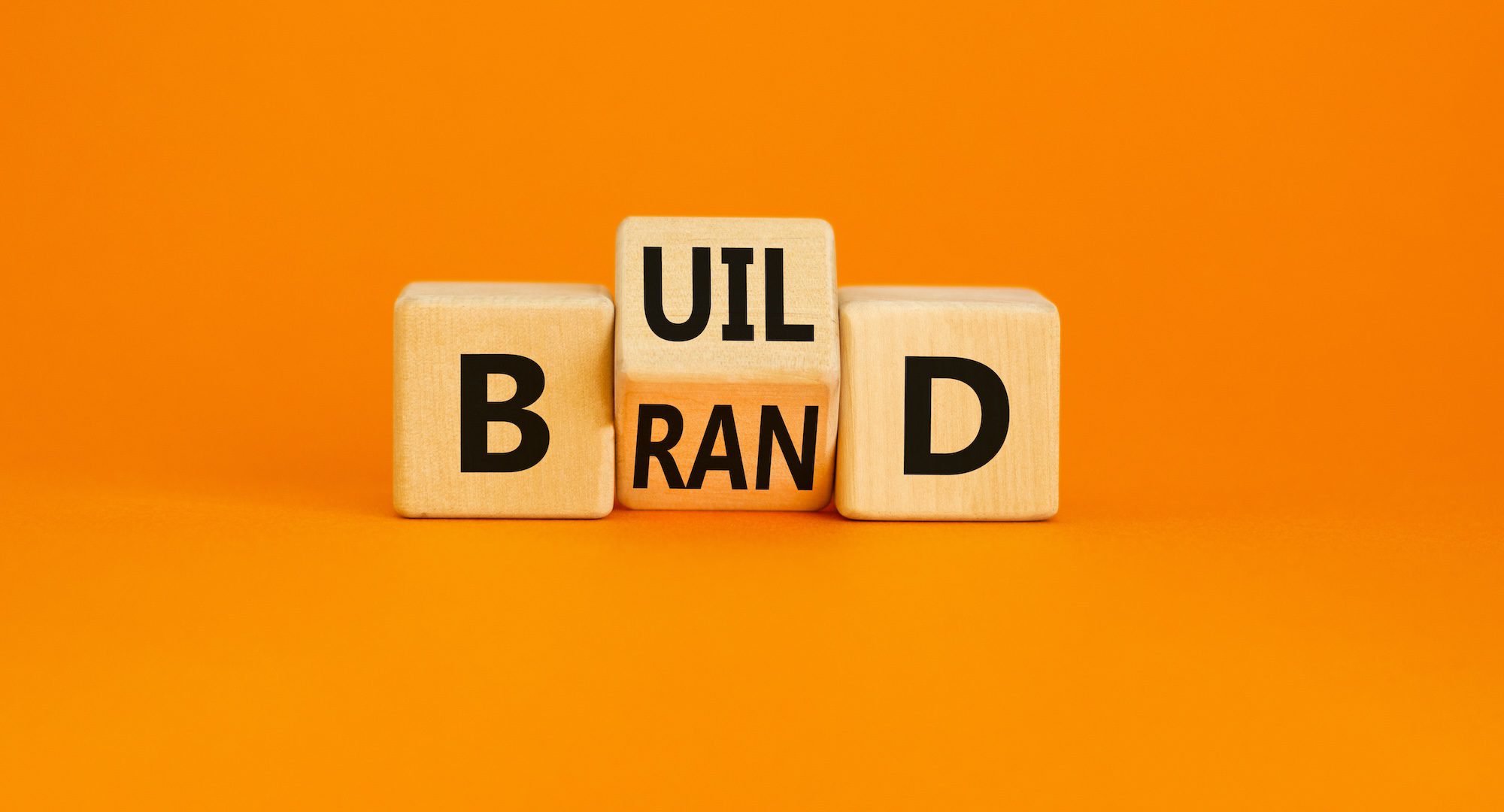 Wooden blocks on an orange background spell "BUILD BRAND," with letters on three separate blocks.