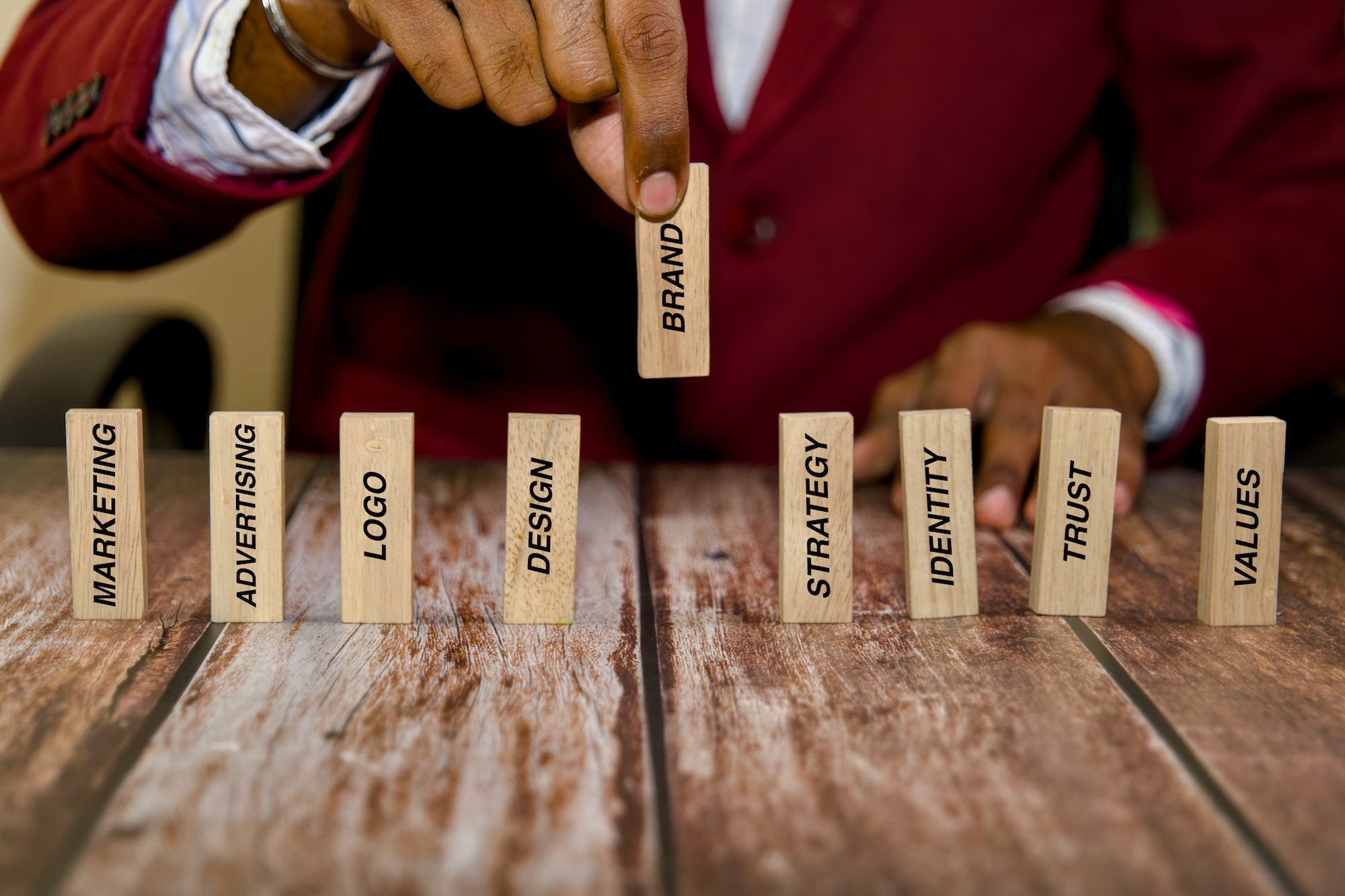 A person in a red suit is arranging wooden blocks on a table. The blocks are labeled with words: "MARKETING," "ADVERTISING," "LOGO," "DESIGN," "BRAND," "STRATEGY," "IDENTITY," "TRUST," and "VALUES." The emphasis is on building a brand.