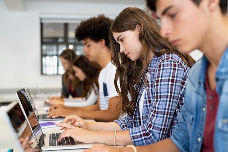 Students sit in a row, focused on typing on their laptops in a classroom setting. They appear engaged in their work, with notebooks and pens nearby. The background shows a window and classroom decor.