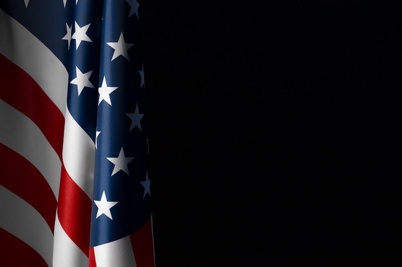 A close-up of the United States flag, showing the stars and stripes on a dark background. The red, white, and blue colors of the flag stand out prominently against the black backdrop.