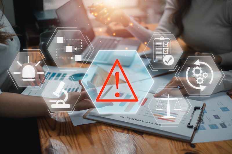 Two people work on a table with documents and charts. Hexagonal icons float above, including an exclamation mark, pie chart, checklist, and scales, symbolizing data analysis, risk assessment, and decision-making.