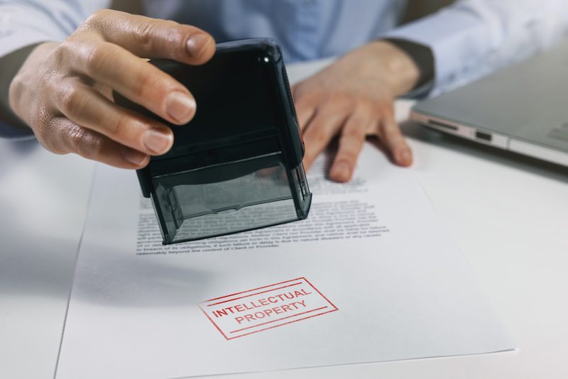 A person stamps a document with "INTELLECTUAL PROPERTY" in red. The document lies next to a laptop on a desk, depicting an official process related to intellectual property rights.