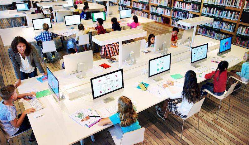 A diverse group of children in a modern library using computers at long desks. A teacher assists a student. Bookshelves line the walls, and each monitor displays educational content. The setting is bright and focused on learning.