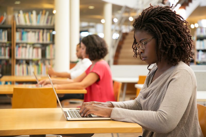 A woman with curly hair and glasses is focused on using a laptop at a library. She is seated at a wooden table, with bookshelves and other people working in the background. The atmosphere is calm and studious.