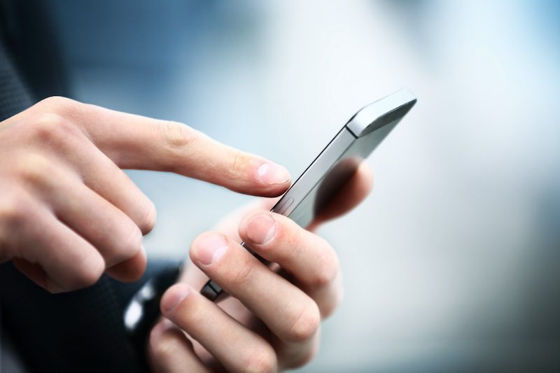 Close-up of a person's hands holding and using a smartphone. One finger is touching the screen, suggesting interaction with the device. The background is blurred, emphasizing focus on the hands and phone.