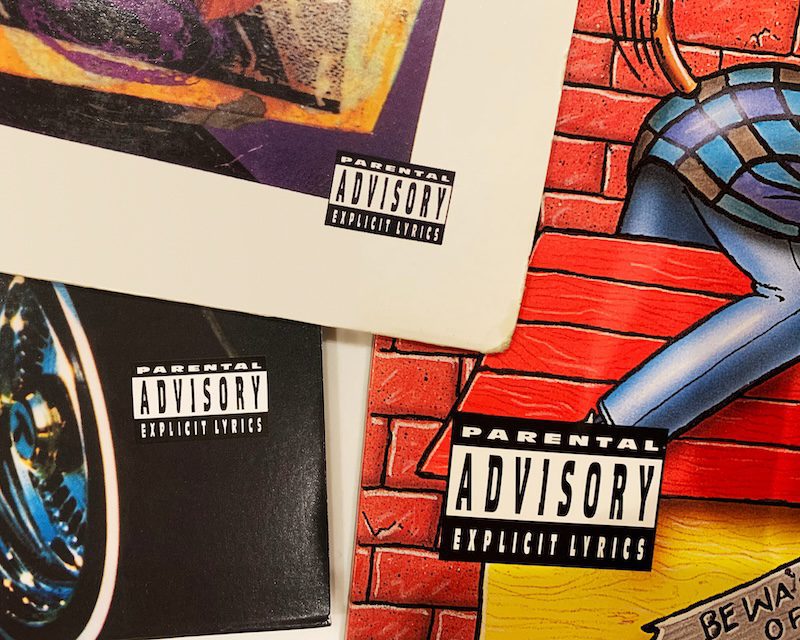 Close-up of three music album covers featuring "Parental Advisory: Explicit Lyrics" labels. The covers display various colorful designs and illustrations, including a cartoon brick wall with blue and purple elements.