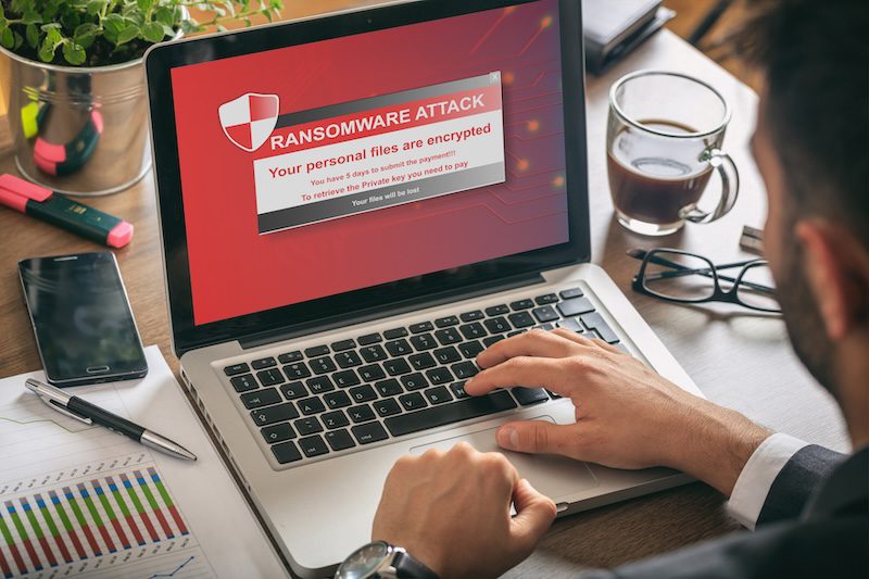 A person sits at a desk with a laptop displaying a ransomware attack warning on the screen. Nearby, there is a smartphone, a cup of coffee, and various documents and charts.