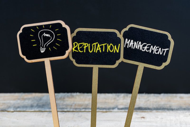 Three small blackboard signs on sticks against a dark background. The first sign shows a lightbulb drawing, the second reads "Reputation," and the third reads "Management." They rest on a wooden surface.