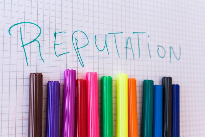 The word "Reputation" is written in blue on graph paper, above a row of colorful markers arranged vertically in various shades, including brown, purple, pink, red, orange, yellow, green, blue, and black.