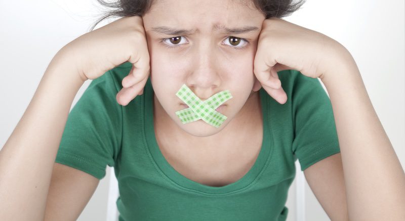 Young person wearing a green shirt, with a sad expression, sits with their head resting on their fists. They have two crossed pieces of green plaid tape over their mouth. The background is light-colored.