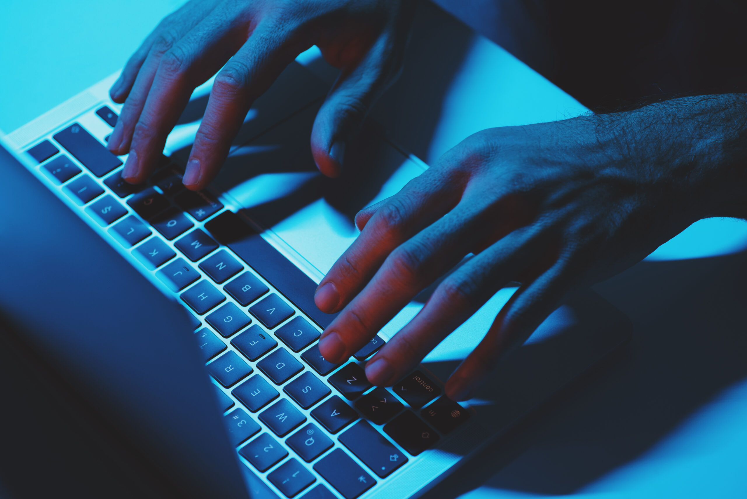 Hands typing on a laptop keyboard, illuminated by blue light.