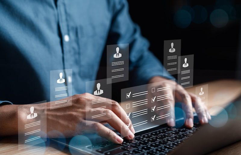 A person typing on a laptop with digital icons of profiles, resumes, and checklists floating above the keyboard. The atmosphere suggests a focus on recruitment or data management.