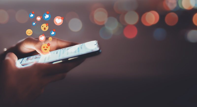 A person holding a smartphone with colorful social media reaction icons, like hearts and thumbs up, floating above the screen. The background is blurred with warm bokeh lights.