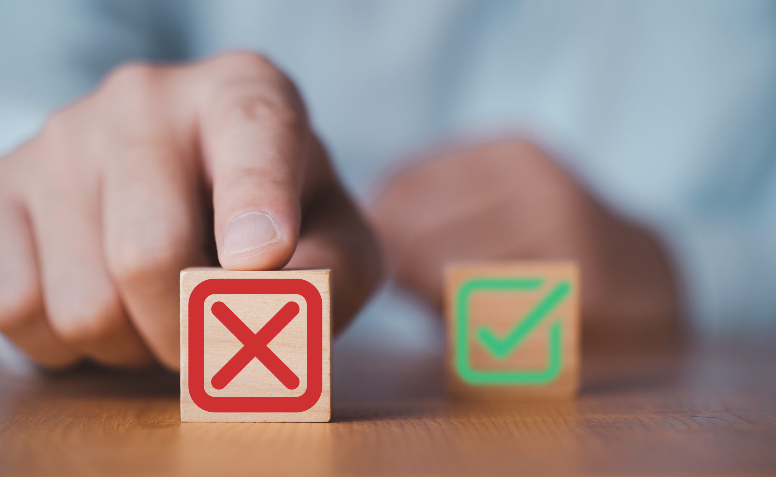 A hand points to a wooden block with a red X symbol. Next to it is another block with a green check mark. The blocks are on a wooden surface, and the background is blurred.