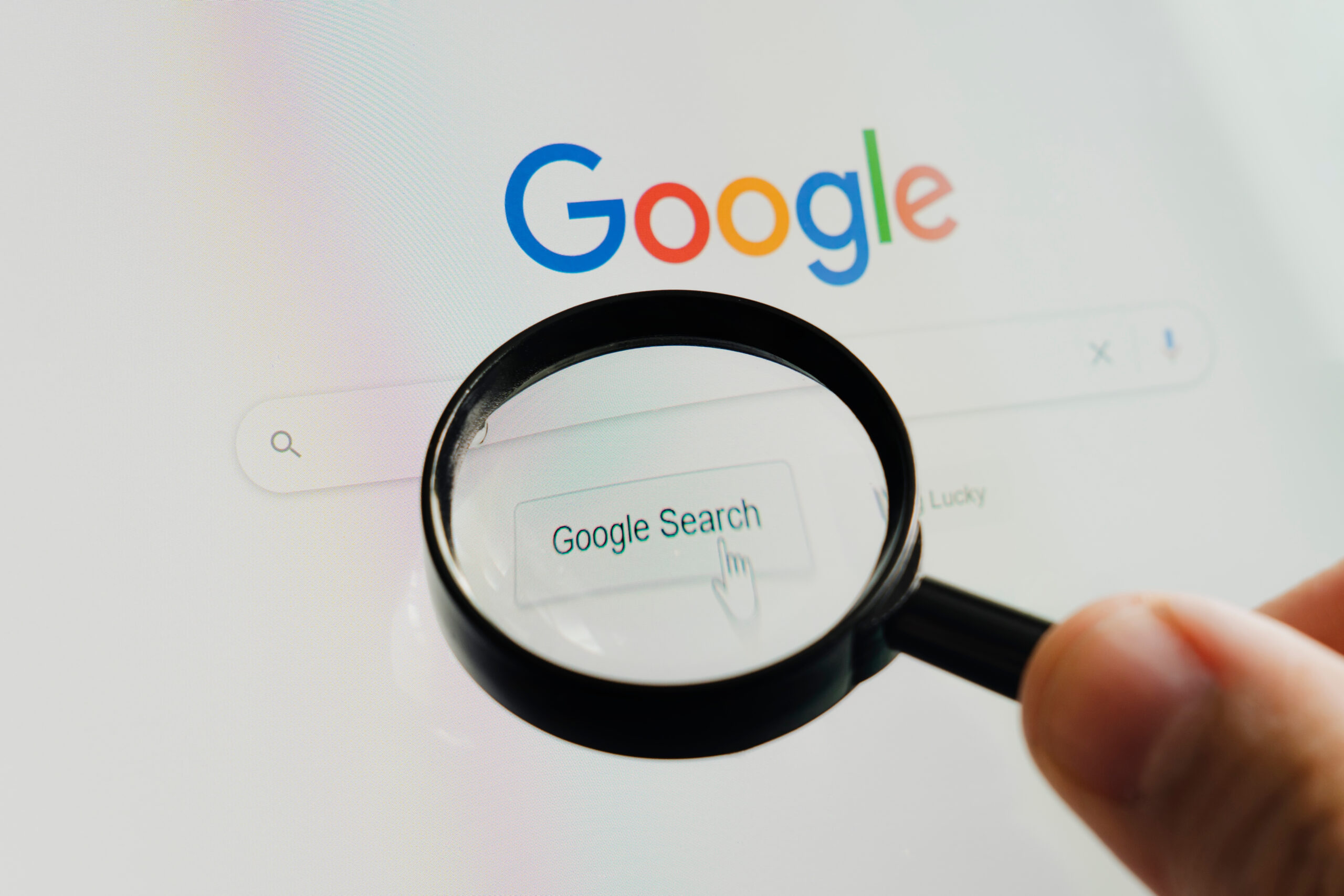 Close-up of a hand holding a magnifying glass over a computer screen, highlighting the Google logo and search bar. The cursor is pointed at the "Google Search" button.