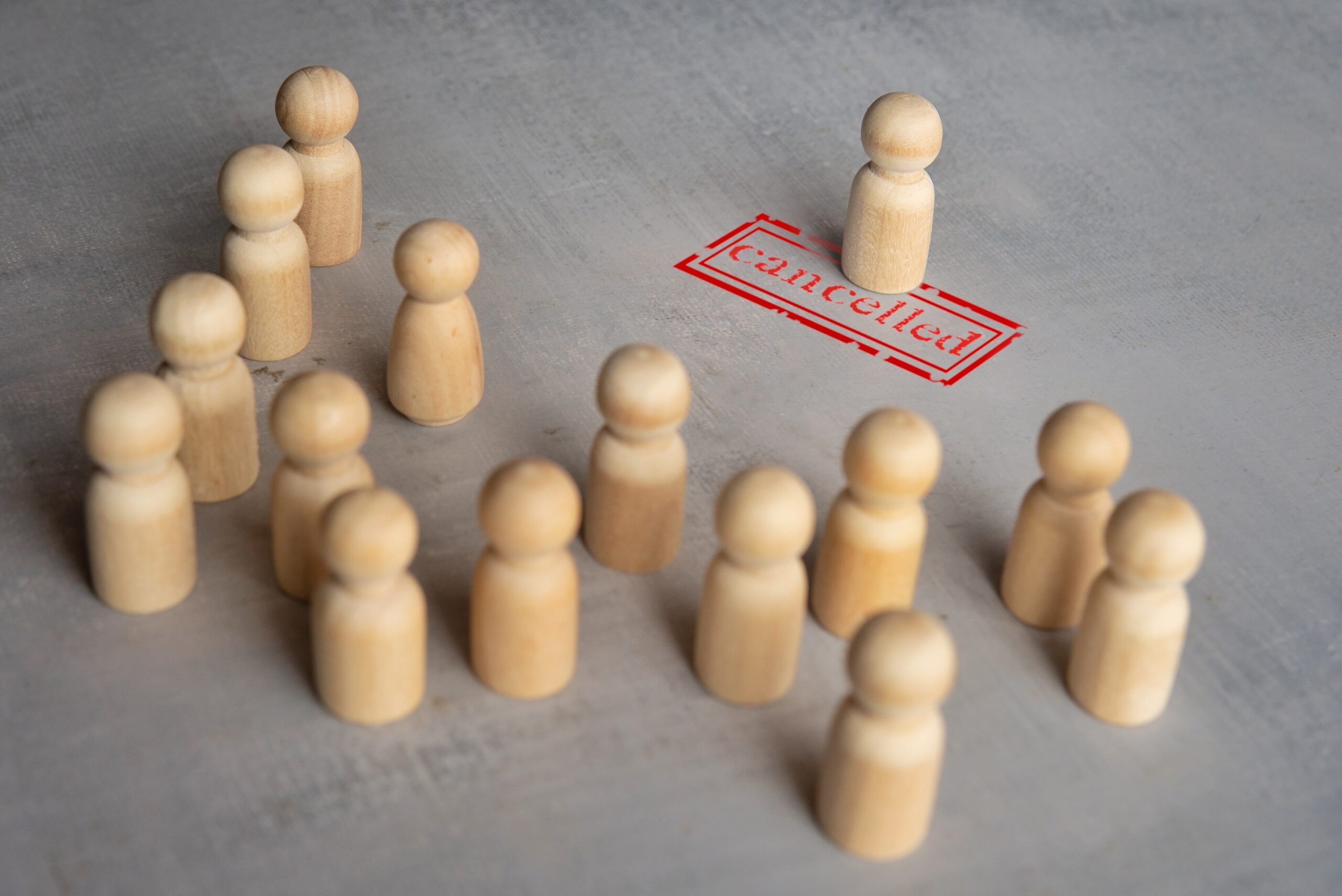 Wooden figurines on a gray surface, with one standing alone next to a red "canceled" stamp, while others are grouped together, suggesting exclusion or separation.